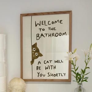 以下が含まれることがあります： 「WELCOME TO THE BATHROOM」と「A CAT WILL BE WITH YOU SHORTLY」のテキストが入ったフレームプリント。漫画の猫がテキストの後ろから覗いています。プリントは、トイレットペーパー、キャンドル、像が置かれた棚の上に表示されています。