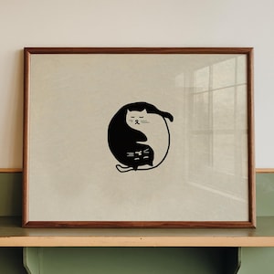 Könnte beinhalten: Ein gerahmter Druck mit einem schwarzen Katzendesign im Yin-Yang-Stil. Das Kunstwerk wird auf einem grünen und beigen Metallregal ausgestellt. Der Rahmen hat eine warme braune Farbe, und der Hintergrund des Drucks ist hellbeige.