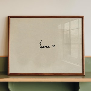 Può includere: Una stampa incorniciata con la parola "home" in corsivo e un piccolo cuore. La cornice è marrone e la stampa è esposta su una mensola verde e color legno montata su una parete bianca. La mensola ha un design arrotondato.