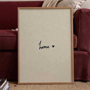 May include: A framed print featuring the word "Home" in black script with a heart symbol. The print has a light beige background and a wooden frame. It is propped against a burgundy sofa.