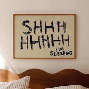 Puede incluir: Un estampado enmarcado con las palabras "SHHH HHHHH I'M SLEEPING" en letras negras, estilo manuscrito. El estampado tiene un fondo beige claro y un marco de madera marrón. La imagen está encima de una cama con almohadas a rayas.
