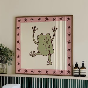 May include: A square framed wall art print featuring a light green cartoon frog with its arms raised and one leg lifted. The illustration is centred on a cream background, surrounded by a pink border decorated with dark brown star shapes.