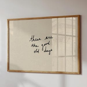 Könnte beinhalten: Ein gerahmter Druck mit dem handgeschriebenen Text "these are the good old days" in schwarzer Schrift. Der Druck hat einen beigefarbenen Hintergrund und ist in einem hellbraunen Holzrahmen eingefasst. Das Bild hängt an einer weißen Wand.