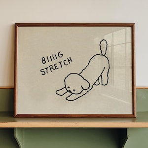 Pode incluir: Uma obra de arte emoldurada com um desenho a traço preto de um cão a alongar-se, com o texto "BIG STRETCH". A obra é exibida numa prateleira verde e de madeira, montada numa parede branca.
