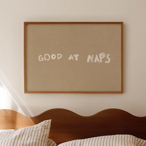 May include: A framed print with the words "GOOD AT NAPS" in white lettering hangs above a bed. The bed has a dark wooden headboard and is covered with a striped duvet and pillows.