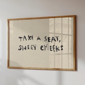 Puede incluir: Una impresión enmarcada con el texto "TAKE A SEAT, SWEET CHEEKS" en fuente manuscrita negra sobre un fondo beige. El marco es de madera marrón claro y la impresión se muestra en una pared blanca.
