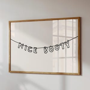 May include: A framed print featuring the words "NICE BOOTY" in black, suspended from a black string. The letters are outlined in black. The frame is made of light brown wood, set against a white background.