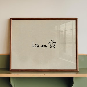 May include: A framed print with the words "bite me" and a gingerbread man illustration. The frame is brown, and the print is displayed on a green and beige shelf attached to a white wall.
