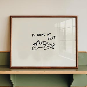 Può includere: Un'opera d'arte incorniciata con la scritta "I'M DOING MY BEST" e un'illustrazione minimalista di una persona sdraiata. L'opera è esposta su una mensola verde e color legno, montata su una parete bianca.