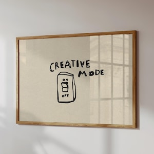 Op de afbeelding: Een ingelijst kunstwerk aan een muur met de woorden "CREATIVE MODE" en een handgetekende lichtschakelaar illustratie. De schakelaar staat in de "ON" positie. Het frame is van hout.
