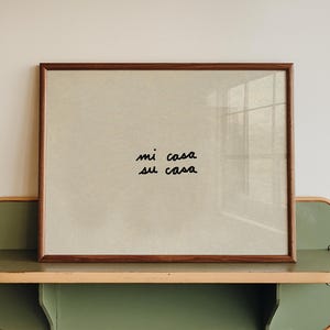 Può includere: Una stampa incorniciata con le parole "mi casa su casa" in grafia nera poggia su una mensola verde e beige. La cornice è di colore marrone scuro. La mensola è montata su una parete bianca.