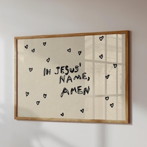 May include: A framed print with the words "IN JESUS' NAME, AMEN" in black handwritten style. Small black heart doodles are scattered across the beige background. The frame is made of light brown wood.