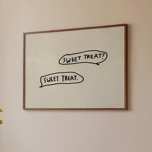 May include: A framed artwork with the text "SWEET TREAT?" and "SWEET TREAT." in black speech bubbles. The artwork hangs above a round wooden table with a vase of yellow flowers and a book. A brown mug sits on the table.