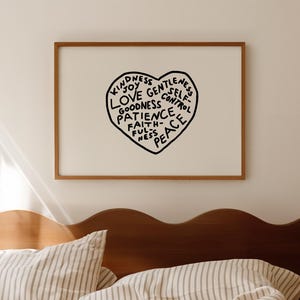 May include: A framed print with a black heart design, inscribed with words such as "Kindness" and "Peace." The artwork is displayed above a bed with striped bedding and a wooden headboard. The room is illuminated by natural light.