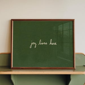 Pode incluir: Uma impressão emoldurada com as palavras "joy lives here" em escrita cursiva sobre um fundo verde. A moldura é marrom e a impressão repousa sobre uma prateleira de dois níveis, verde e bege, fixada a uma parede branca.