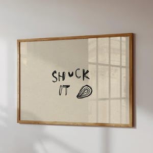 Może przedstawiać: Oprawiony wydruk z napisem "SHUCK IT" czarną, odręczną czcionką, wraz z czarną ilustracją ostrygi. Grafika ma beżowe tło i ramę z naturalnego drewna, zamontowaną na białej ścianie.