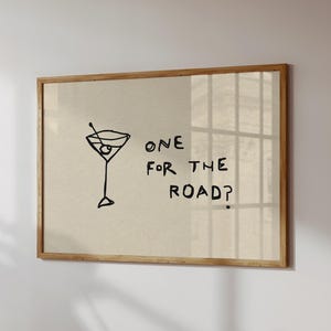 以下が含まれることがあります： 黒い線で描かれたマティーニグラスのシンプルなイラストと「ONE FOR THE ROAD？」の文字がベージュの背景に描かれた額入りのプリント。木製のフレームに入っており、白い壁に飾られています。