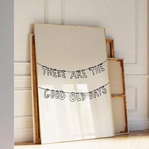 Pode incluir: Uma impressão em tela emoldurada com o texto "THESE ARE THE GOOD OLD DAYS" em escrita preta. A impressão tem uma moldura dourada e está encostada em uma parede branca. A obra de arte é exibida em uma sala com piso de madeira.