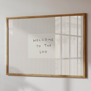 May include: A framed sign with a wooden frame, featuring a white background with vertical grey stripes. The sign includes a white paper with the text "WELCOME TO THE LOO" in black ink.