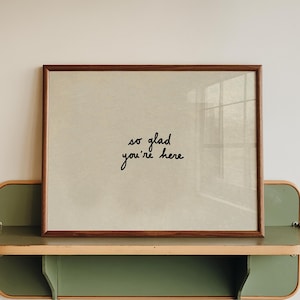 May include: A framed print with the text "so glad you're here" sits on a green and wood-toned shelf. The frame is a dark brown colour, and the shelf has a rounded design. The background is a plain white wall.