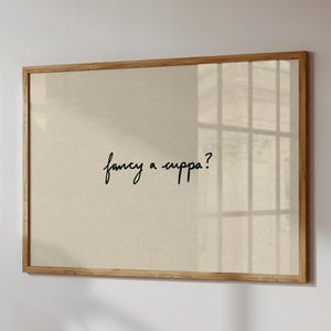 Puede incluir: Un cuadro enmarcado con un fondo beige claro y la frase manuscrita "fancy a cuppa?" en escritura negra. El marco es de madera clara. La impresión está colgada en una pared blanca.