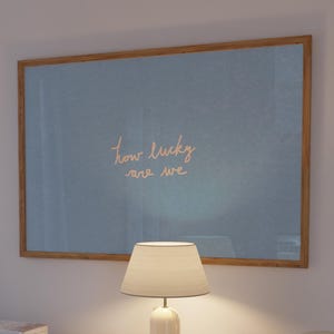 Puede incluir: Una obra de arte enmarcada con el texto "how lucky are we" en escritura dorada cuelga sobre un mueble blanco. Una lámpara blanca con una pantalla beige está sobre el mueble, junto a una pila de libros. La obra tiene un fondo azul claro.