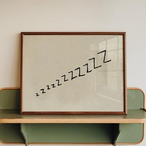 Peut inclure: Une œuvre d'art encadrée avec le mot "zzzzzzzz" en noir sur un fond beige. L'œuvre est exposée sur une étagère verte et boisée, fixée à un mur blanc. L'étagère a un design arrondi.
