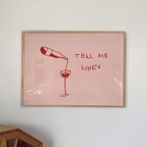 May include: A framed print with a pink background and red illustration of a wine bottle pouring into a glass. The words "TELL ME WHEN" are written in red. The frame is a light brown color.
