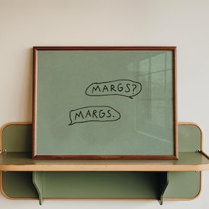 May include: A framed artwork with the text "MARGS?" and "MARGS." in speech bubbles. The artwork is displayed on a green and wood-toned shelf, mounted on a white wall. The frame is a dark brown color.