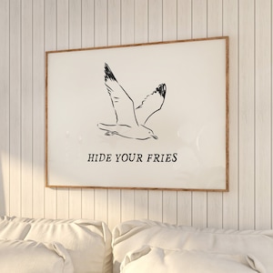 Könnte beinhalten: Ein gerahmter Druck mit einer Schwarz-Weiß-Illustration einer fliegenden Möwe. Der Text "HIDE YOUR FRIES" steht darunter. Das Kunstwerk wird an einer weißen Wand mit Paneelen über einem weißen Sofa mit Kissen gezeigt.