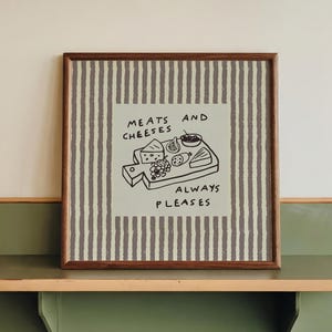 Pode incluir: Uma impressão emoldurada com um fundo listrado e as palavras "MEATS AND CHEESES ALWAYS PLEASES" acima de um desenho de uma tábua de cortar com comida. A impressão é exibida em uma prateleira verde e bege.