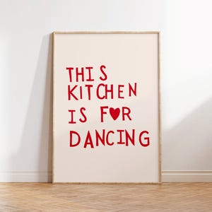 以下が含まれることがあります： 赤色の太字で「THIS KITCHEN IS FOR DANCING」と書かれた額入りのプリント。プリントはクリーム色の背景と木製のフレームです。プリントは、木製の床の白い壁に立てかけられています。