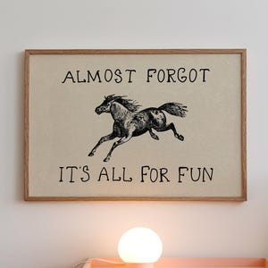 May include: A framed print with the text "ALMOST FORGOT IT'S ALL FOR FUN" and a black and white illustration of a running horse. The artwork is in a wooden frame and hangs on a white wall above a pink nightstand.