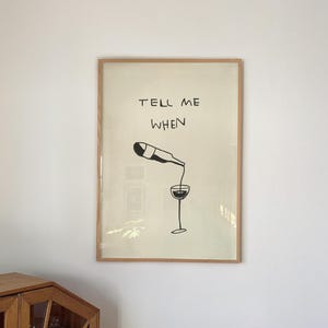 Puede incluir: Un cuadro enmarcado con una ilustración minimalista en blanco y negro de una botella de vino vertiendo en una copa. El texto "TELL ME WHEN" está encima de la ilustración. El marco es de color marrón claro.