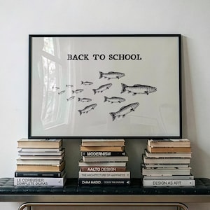 May include: A framed horizontal print featuring the text BACK TO SCHOOL in black capital letters above a black and white illustration of a school of fish swimming in a group. The print is displayed on three stacks of books on a black bench.