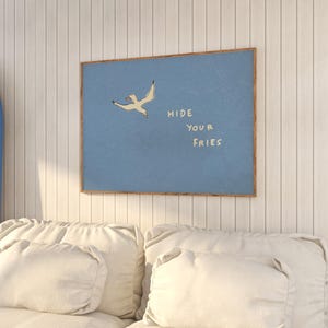 May include: A framed art print featuring a light blue background with a seagull illustration and the text "HIDE YOUR FRIES" in white. The artwork is displayed on a white paneled wall above a white sofa.