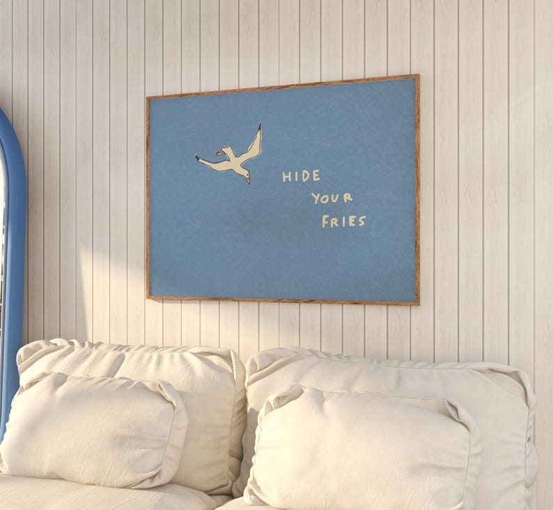 May include: A framed art print featuring a light blue background with a seagull illustration and the text "HIDE YOUR FRIES" in white. The artwork is displayed on a white paneled wall above a white sofa.