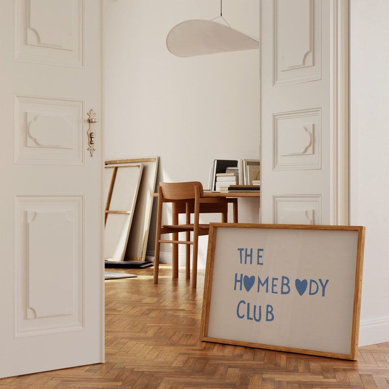 May include: A framed print with a light brown background and a blue inscription that reads "THE HOMEBODY CLUB" with two blue hearts.