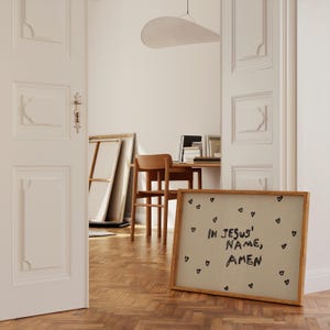 May include: A framed artwork with the text "IN JESUS' NAME, AMEN" in black script, surrounded by small black heart doodles. The frame is a light brown color. The artwork is displayed on a wooden floor.