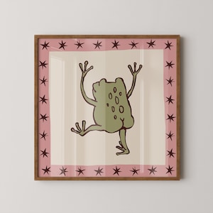 May include: A square framed wall art piece featuring a central illustration of a green frog with dark brown outlines on a cream background. The frog is depicted from behind with arms raised. A pink border surrounds the centre, decorated with dark brown stars.