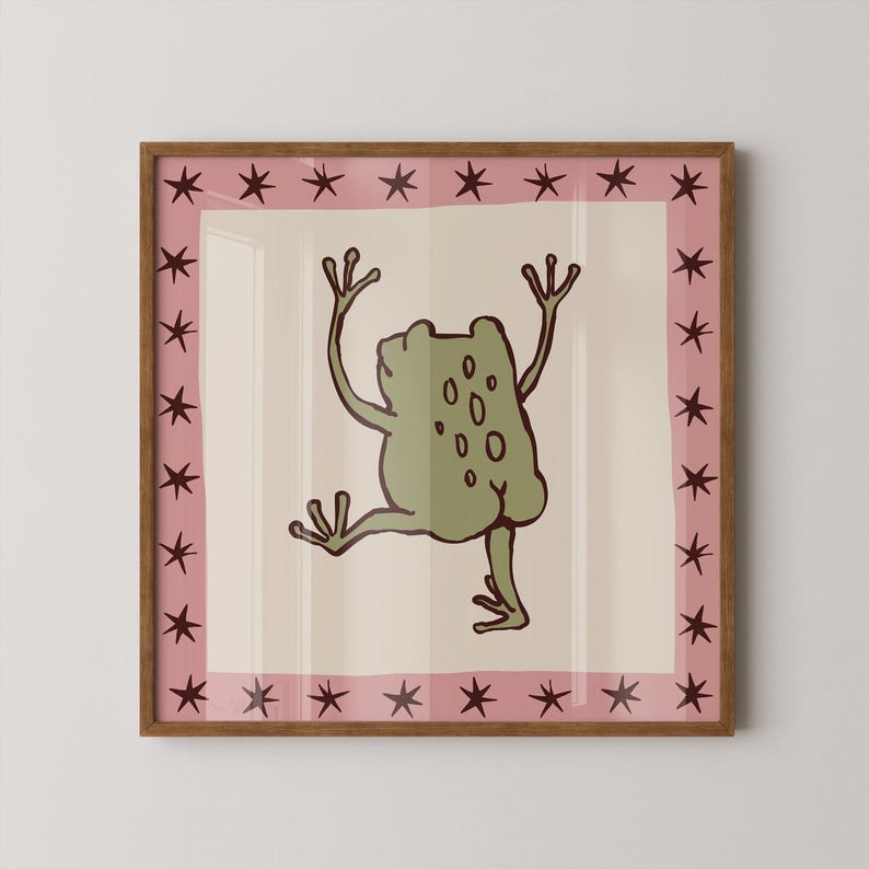 May include: A square framed wall art piece featuring a central illustration of a green frog with dark brown outlines on a cream background. The frog is depicted from behind with arms raised. A pink border surrounds the centre, decorated with dark brown stars.