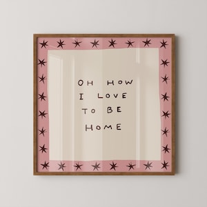 May include: A square framed wall art piece featuring a light beige centre with the dark brown text "OH HOW I LOVE TO BE HOME". The design is surrounded by a pink border decorated with a repeating pattern of dark brown hand-drawn star shapes.