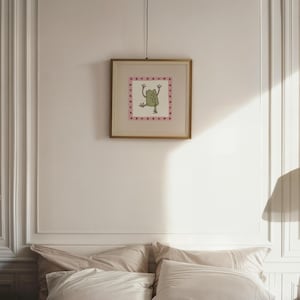 May include: A square framed wall art piece featuring a central illustration of a green frog with raised arms. The frog is enclosed within a pink border decorated with small dark red star shapes. The artwork is encased in a thin gold-toned frame.