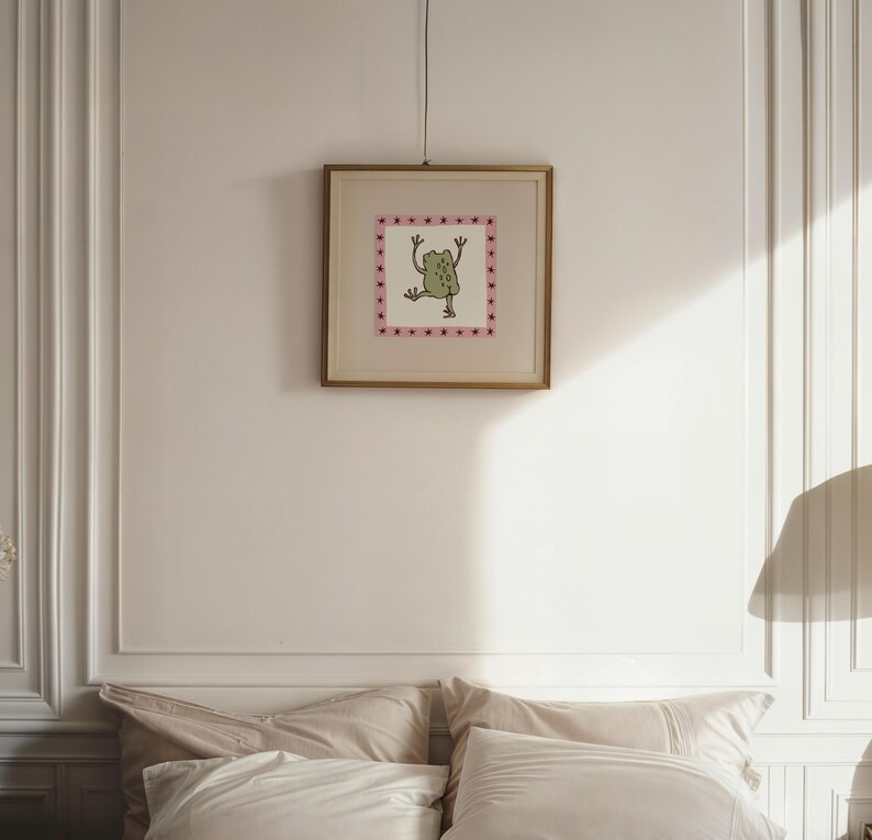 May include: A square framed wall art piece featuring a central illustration of a green frog with raised arms. The frog is enclosed within a pink border decorated with small dark red star shapes. The artwork is encased in a thin gold-toned frame.