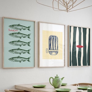 Puede incluir: Tres impresiones enmarcadas en una pared blanca. La impresión de la izquierda tiene un fondo azul claro con una fila de cinco ilustraciones en blanco y negro de sardinas y la palabra "Sardinas" en rojo. La impresión del medio tiene un fondo a cuadros amarillo y blanco con una ilustración azul y blanca de una lata de sardinas. La impresión de la derecha tiene un fondo a rayas verde oscuro y blanco con una ilustración de pez rojo.