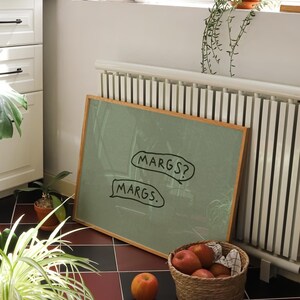 May include: A framed green chalkboard with the text "MARGS?" and "MARGS." in speech bubbles. A woven basket filled with apples sits on a checkered floor. A cookbook and potted plants are also visible.