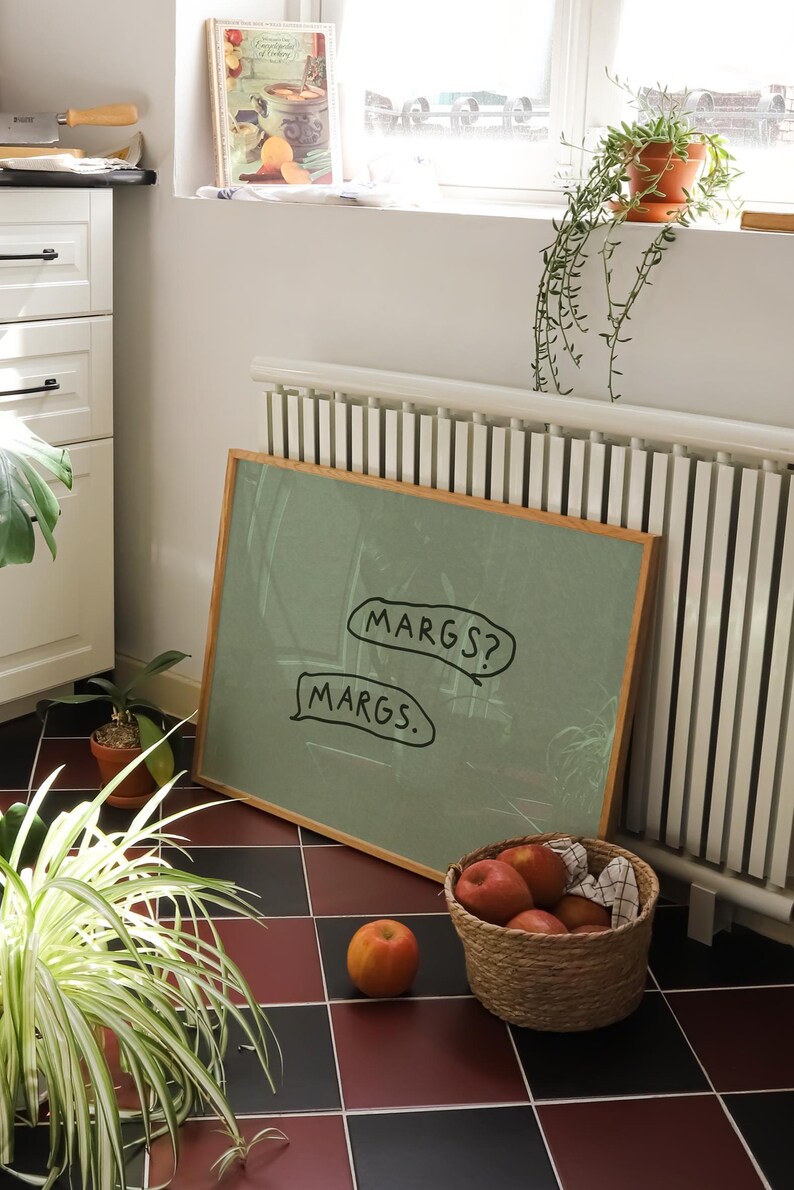 May include: A framed green chalkboard with the text "MARGS?" and "MARGS." in speech bubbles. A woven basket filled with apples sits on a checkered floor. A cookbook and potted plants are also visible.