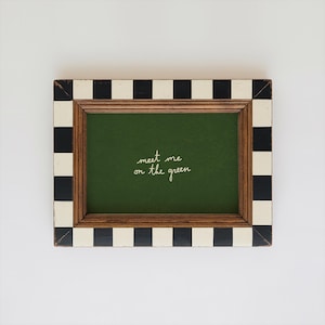 May include: Rectangular picture frame with a green interior and the handwritten text "meet me on the green." The frame has a wooden border and a black and white checkered pattern on the outer edges.