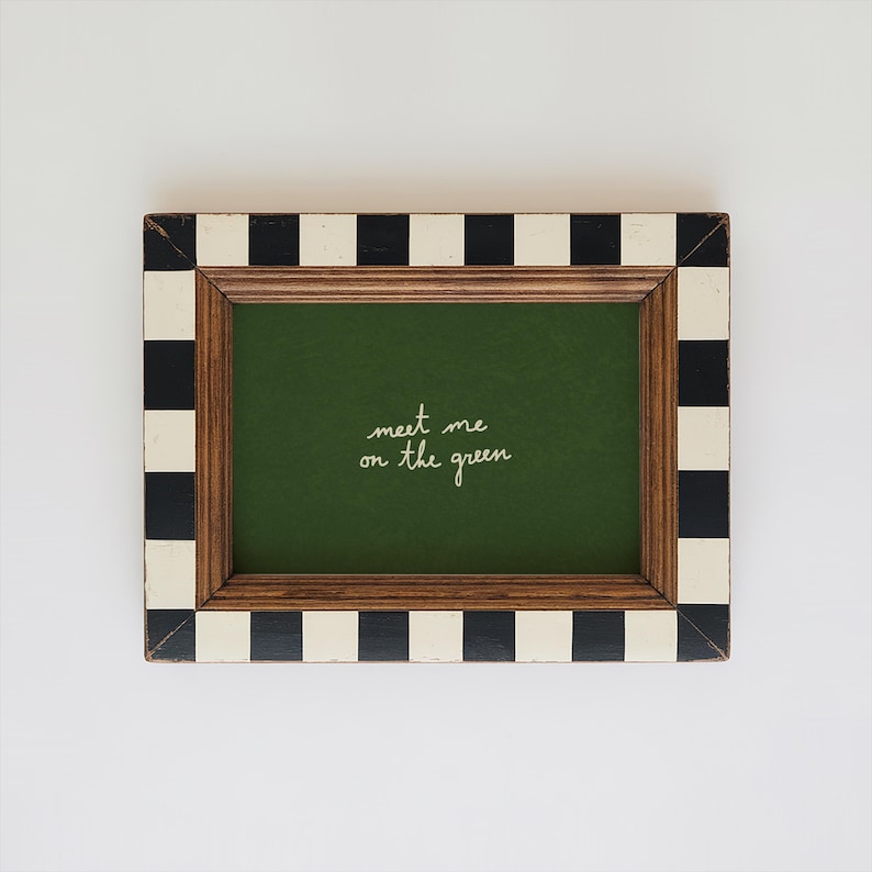 May include: Rectangular picture frame with a green interior and the handwritten text "meet me on the green." The frame has a wooden border and a black and white checkered pattern on the outer edges.