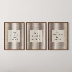 May include: Three rectangular wall art prints in brown wooden frames. Each print features a vertical gray and white striped background with a central white square containing black text. The text reads I SEE LONDON I SEE FRANCE, HEY SWEET CHEEKS, and THE BEST SEAT IN THE HOUSE.